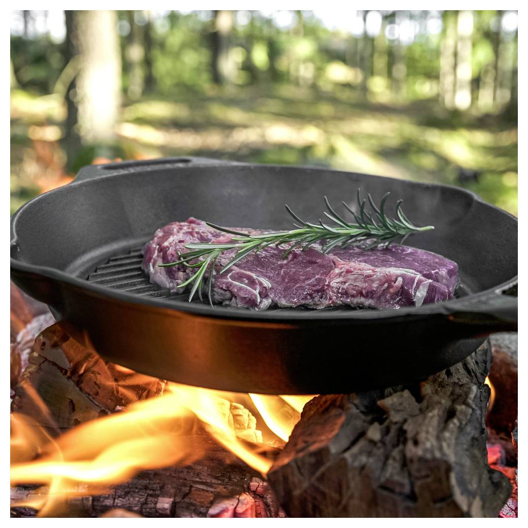 Rohes Steak mit Rosmarin wird in einer Gusseisenpfanne über einem offenen Lagerfeuer in einer waldigen Außenumgebung angebraten.