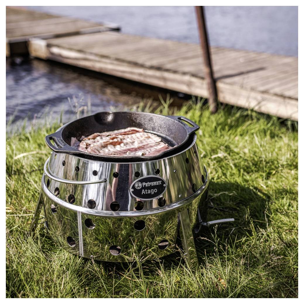 Speck, der in einem tragbaren Edelstahl-Feuerschalen-Grill auf Gras neben einem hölzernen Steg kocht.