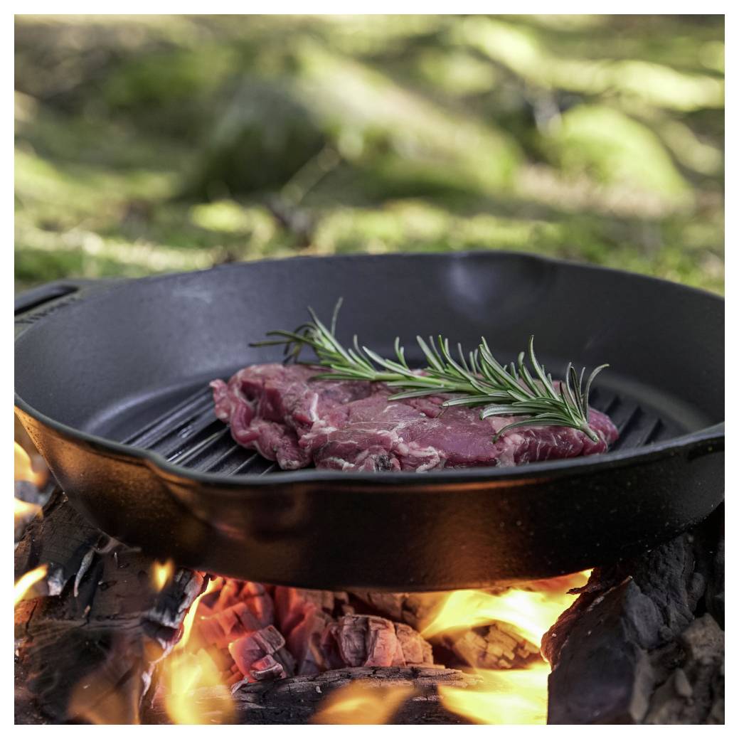 Rohes Steak mit Rosmarin brät in einer gusseisernen Pfanne über einem offenen Holzfeuer im Freien.