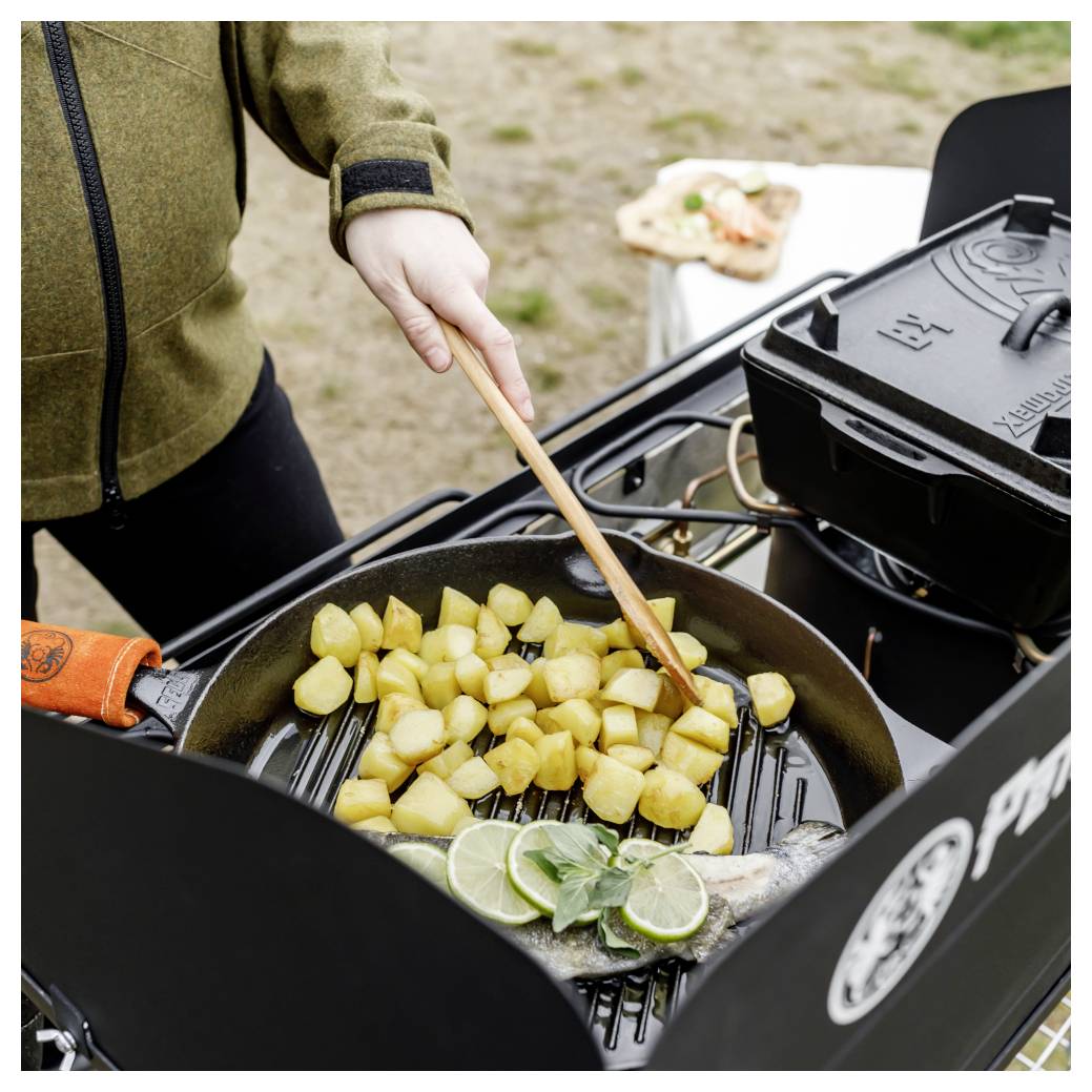 Person kocht Fisch und gewürfelte Kartoffeln in einer Pfanne auf einem Außengrill, mit Limettenscheiben und Kräutern hinzugefügt.