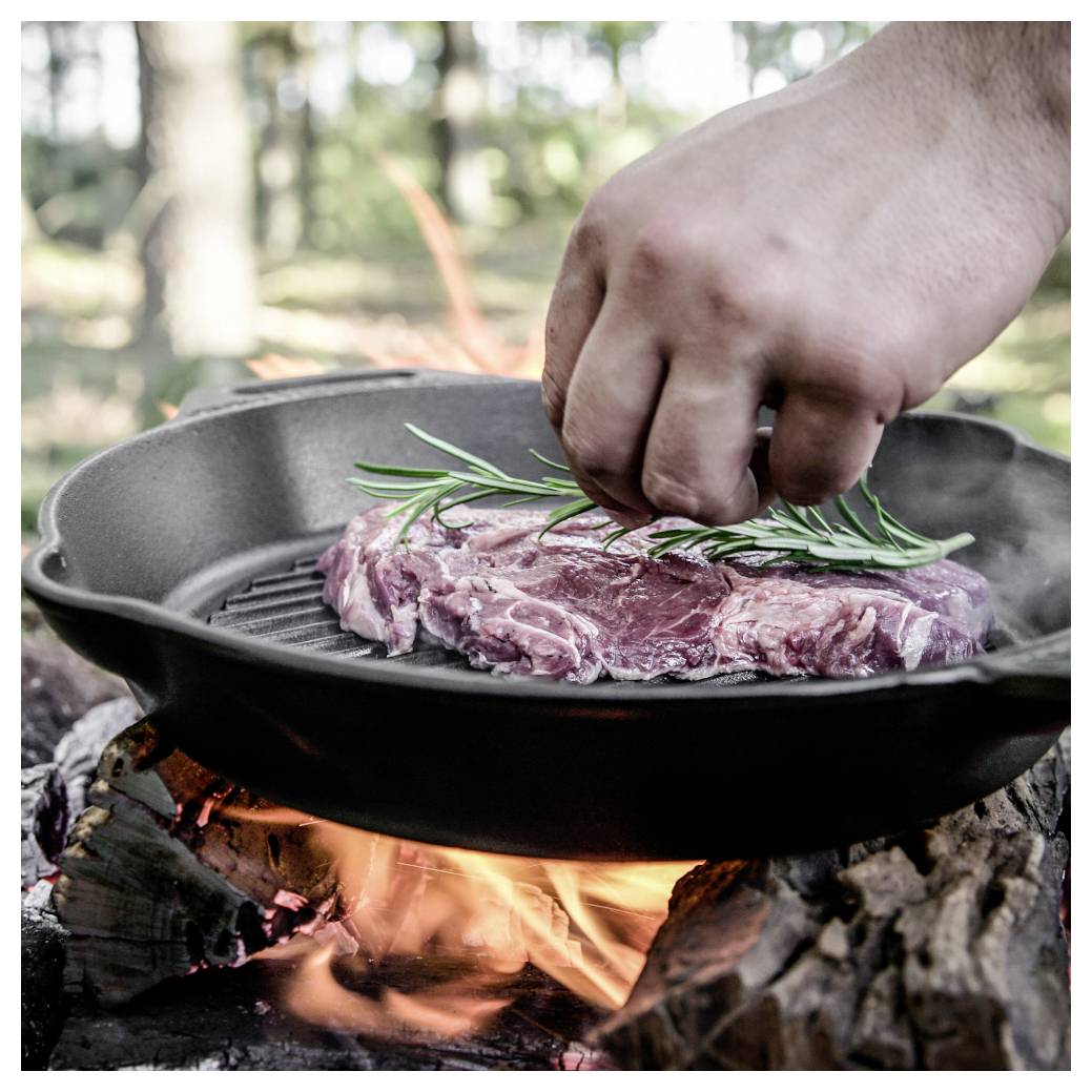Hand würzt ein rohes Steak mit Rosmarin in einer gusseisernen Pfanne über einem offenen Lagerfeuer.
