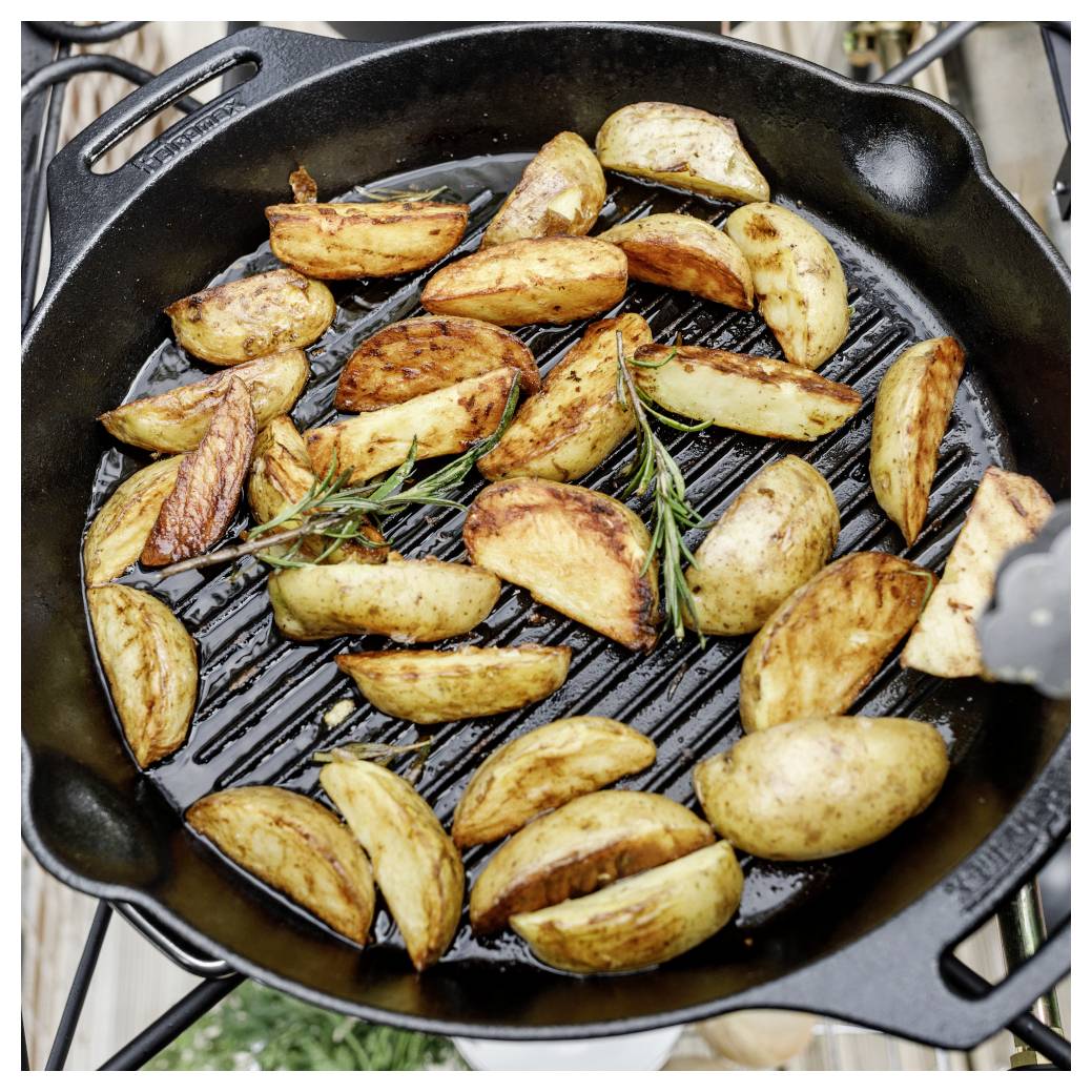 Kartoffelspalten, die in einer gusseisernen Pfanne auf einem Grill rösten, mit Rosmarinzweigen.