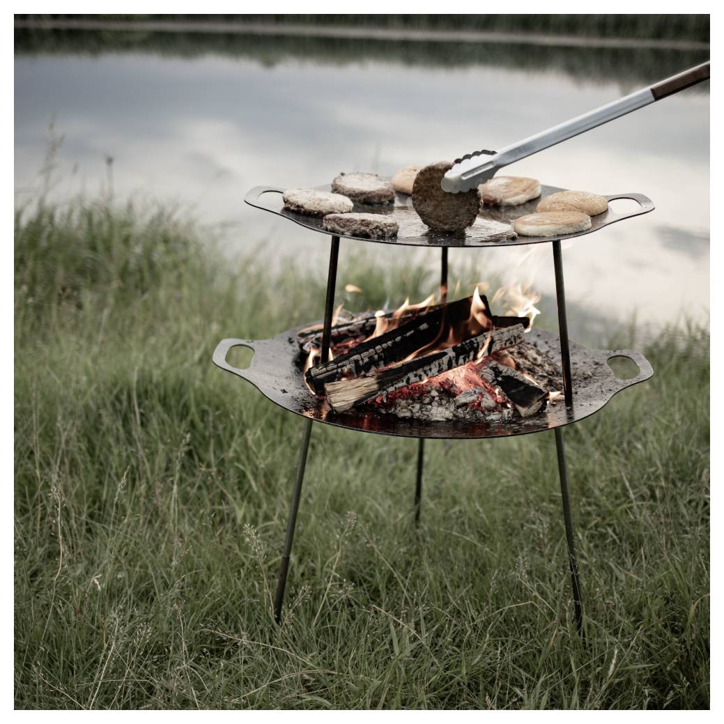 Außengrill mit Burgern und Brötchen, die über einem offenen Lagerfeuer neben einem See zubereitet werden, gezeigt mit Zangen, die ein Patty hochheben.