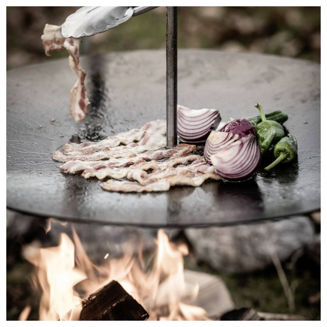 Speck brät auf einem heißen Außengrill über offenem Feuer, mit geschnittenen roten Zwiebeln und grünen Paprikaschoten daneben.