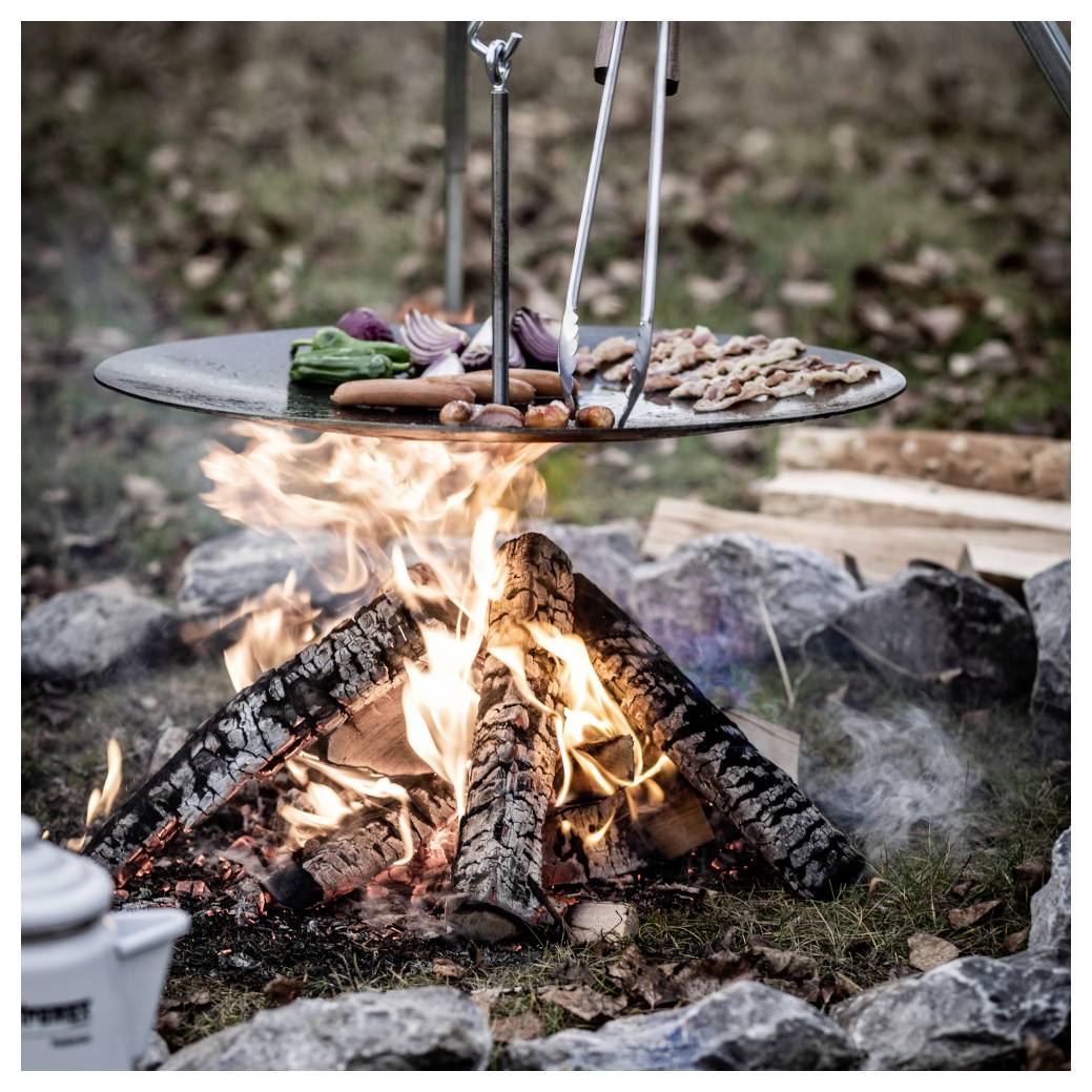 Lebensmittel, die auf einem Metallrost über einem Lagerfeuer garen, mit Gemüse und Fleisch, das über brennenden Holzscheiten brät und von Steinen umgeben ist.