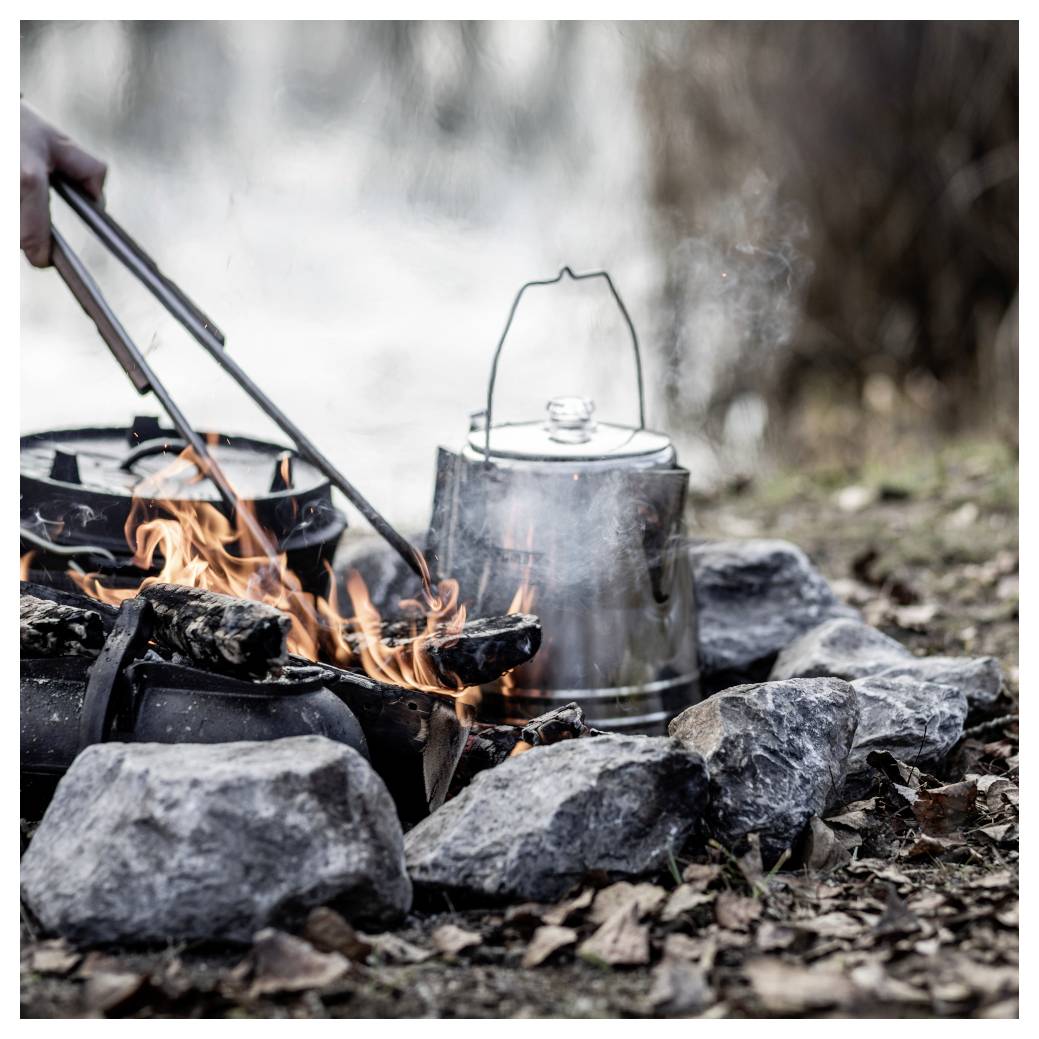 Lagerfeuer mit brennendem Holz und einem Metallkessel, der sich unter Steinen an einem Outdoor-Campingplatz erhitzt.