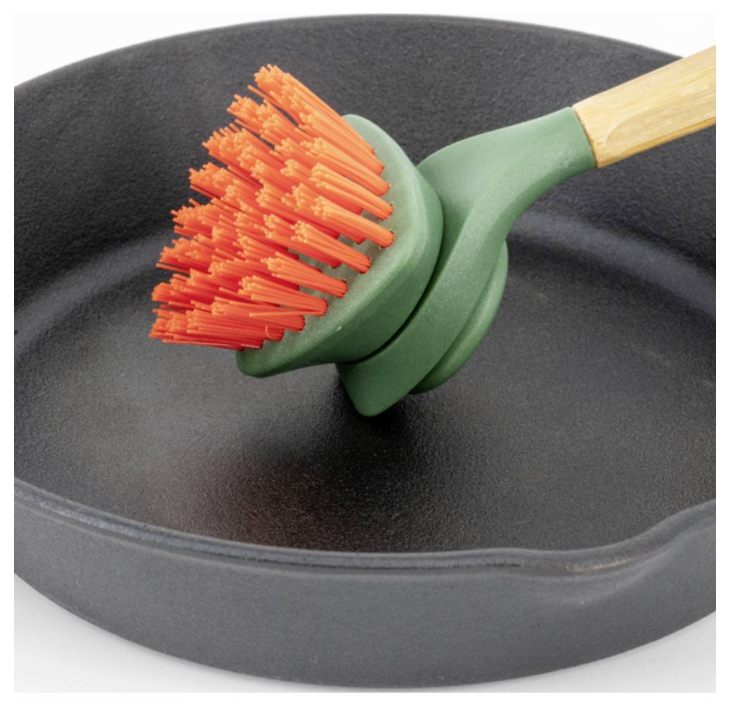 Nahaufnahme einer grünen Spülbürste mit orangefarbenen Borsten, die in einer schwarzen Pfanne ruht, die ein Küchenreinigungswerkzeug zeigt.