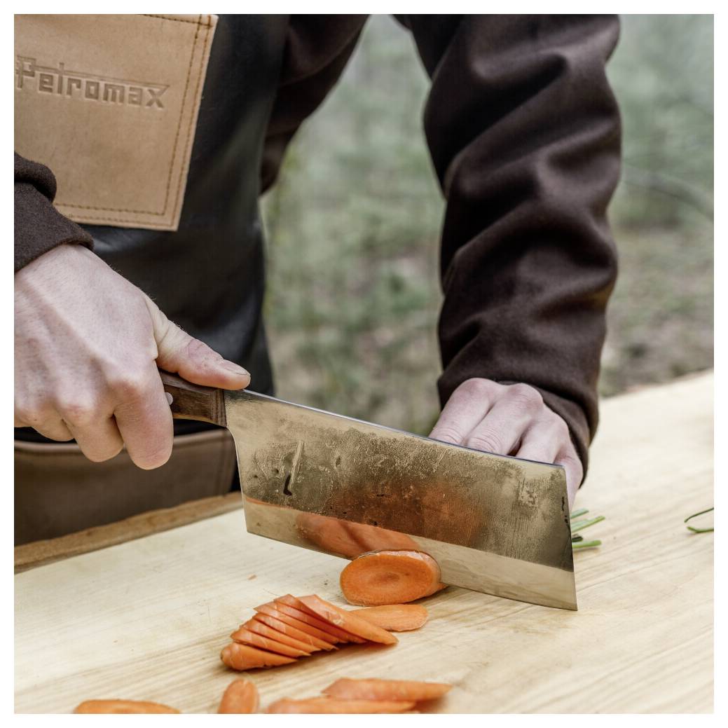 Eine Person schneidet eine Karotte mit einem großen Messer auf einem Holzschneidebrett im Freien.