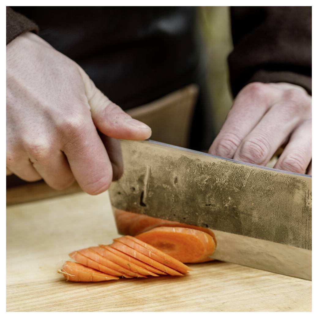 Nahaufnahme von Händen, die eine Möhre mit einem großen Messer auf einem Holzschneidebrett schneiden.