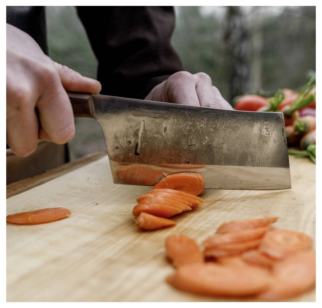 Eine Person schneidet eine Möhre mit einem großen Messer auf einem Holzschneidebrett, mit gehackten Stücken im Vordergrund und unscharfem Gemüse im Hintergrund.