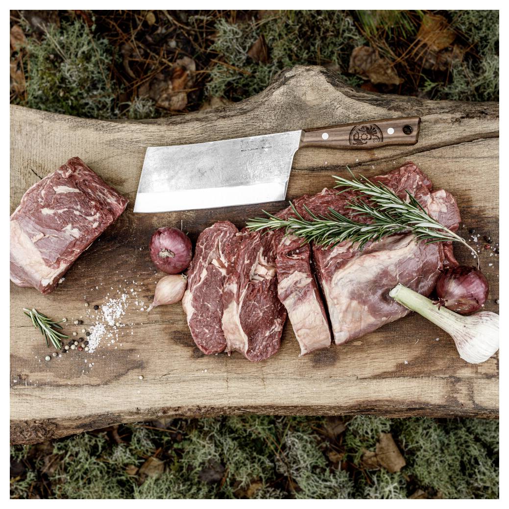 Rohes Rindfleisch Stücke und Gewürze auf einem Holz-Schneidebrett, mit einem Hackebeil und Rosmarinzweigen, arrangiert zur Zubereitung von Outdoor-Speisen.