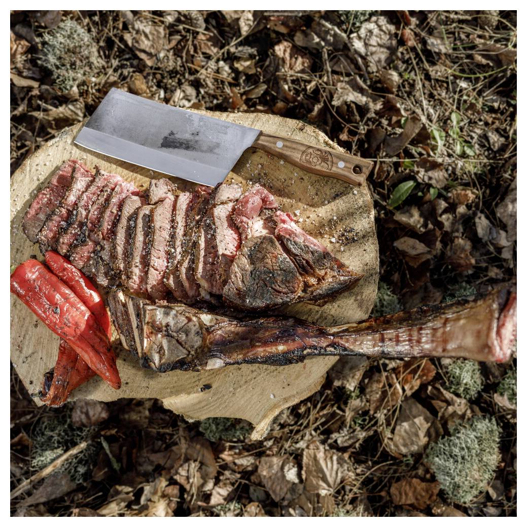 Geschnittenes gekochtes Fleisch auf einem Holzschneidebrett im Freien, mit einem großen Hackebeil und einem roten Utensil daneben.