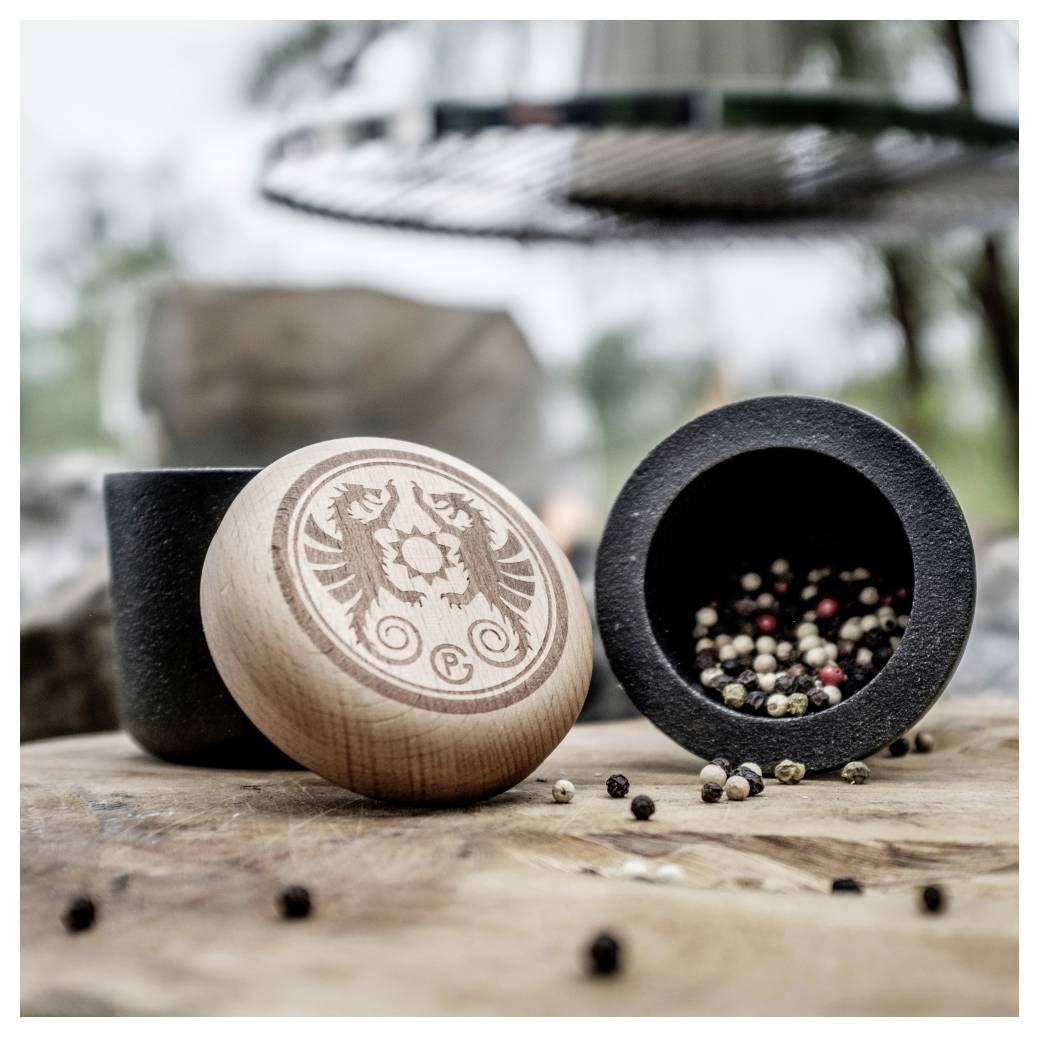 Holzerner Pfeffermühlendeckel und schwarze Gewürzmühle mit gemischten Pfefferkörner, die auf einer rustikalen Oberfläche im Freien verstreut sind.