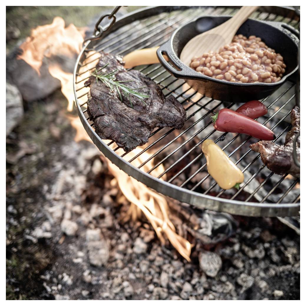 Steak, Bohnen und Paprika kochen auf einem Rost über einem offenen Lagerfeuer, wobei Flammen unter dem Gitter sichtbar sind.