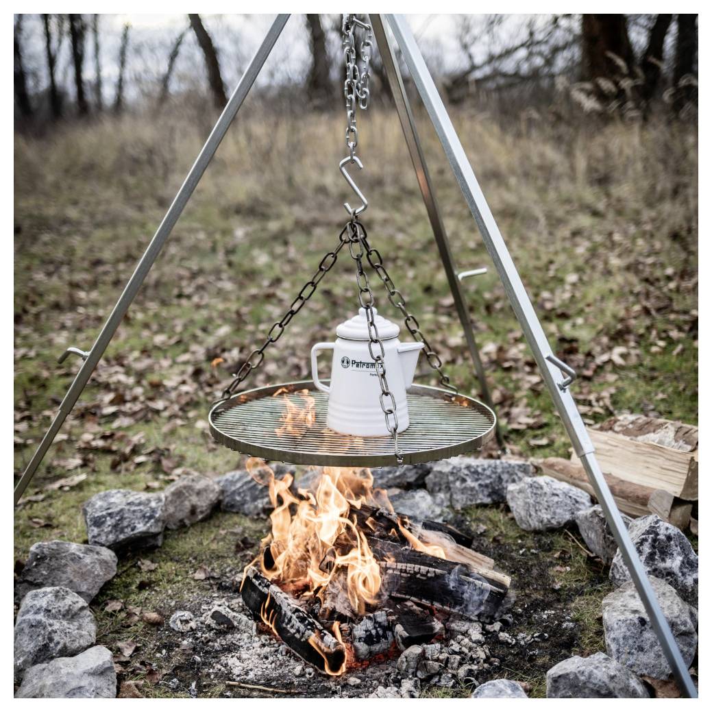 Camping-Dreibein über einem Lagerfeuer mit einem aufgehängten Grillrost und weißem Topf, der über brennenden Holzscheiten in einer Steinfeuerringe kocht.