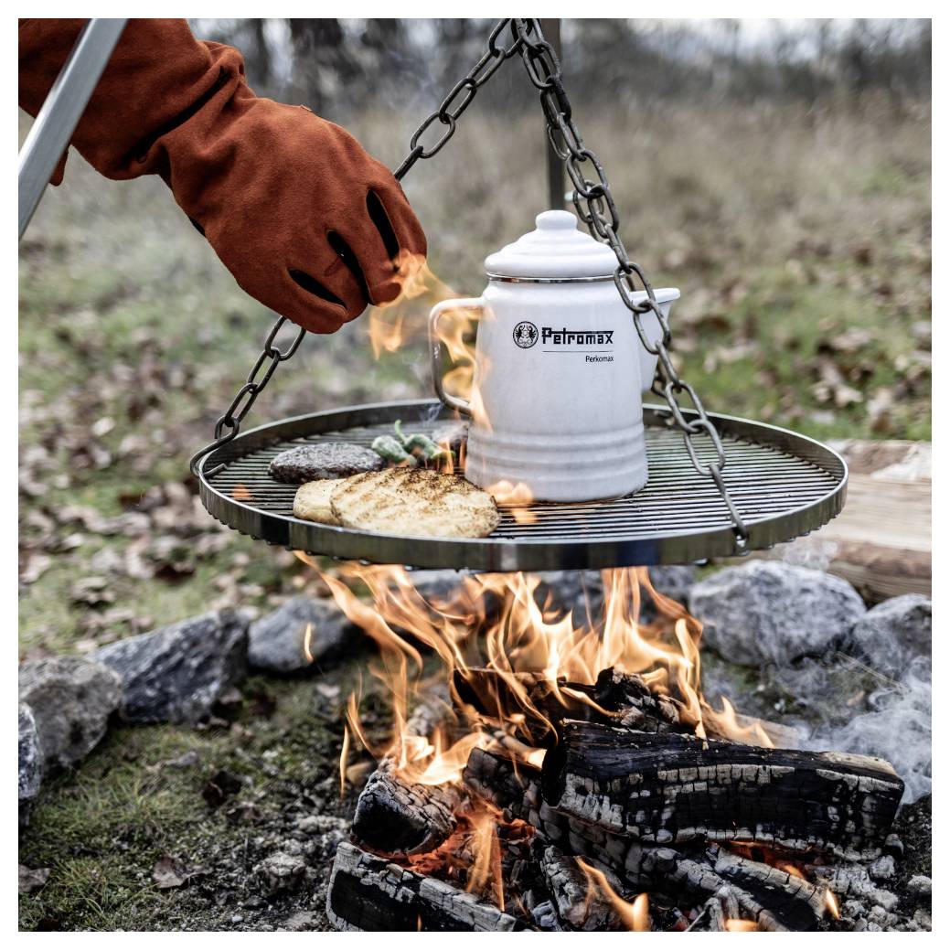 Behandschuhte Hand kocht Essen und erhitzt einen Petromax-Topf auf einem hängenden Grillrost über einem offenen Lagerfeuer im Freien.