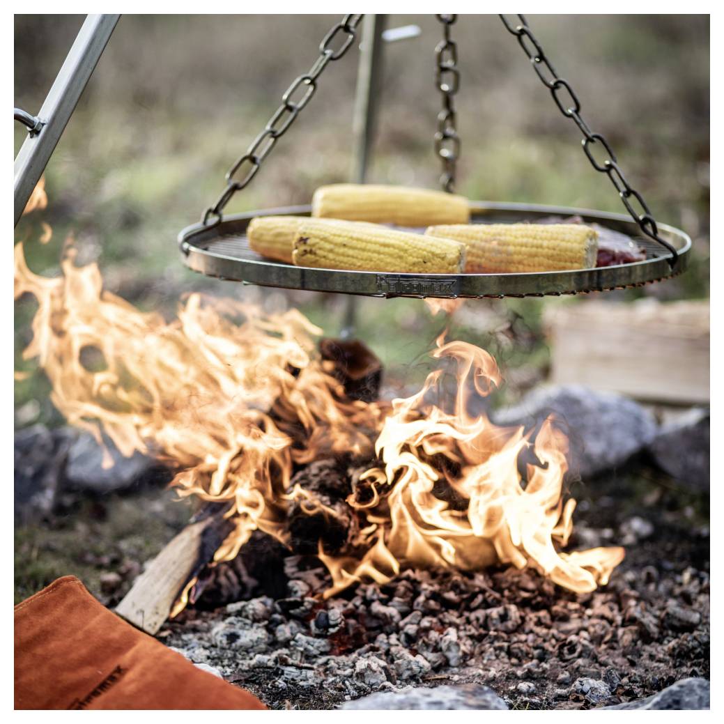 Mais am Kolben ggart auf einem hängenden Rost über einem offenen Lagerfeuer mit Flammen und Glut.