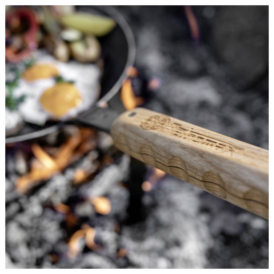 Nahaufnahme einer Bratpfanne über einem offenen Lagerfeuer, mit einem Holzgriff im Vordergrund und Eiern, die darin braten.