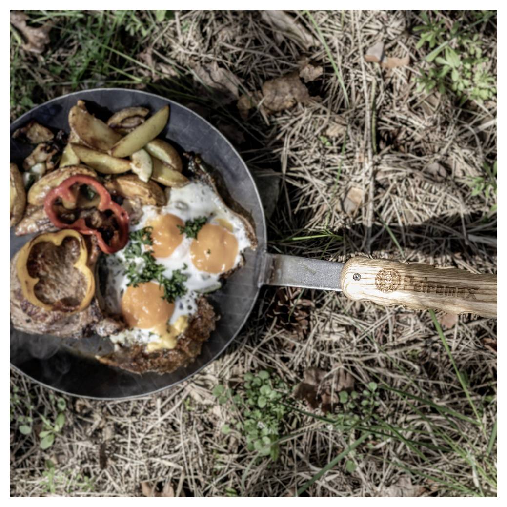 Bratpfanne mit Frühstückseiern, Kartoffelecken und Gemüse auf Gras, von oben betrachtet.