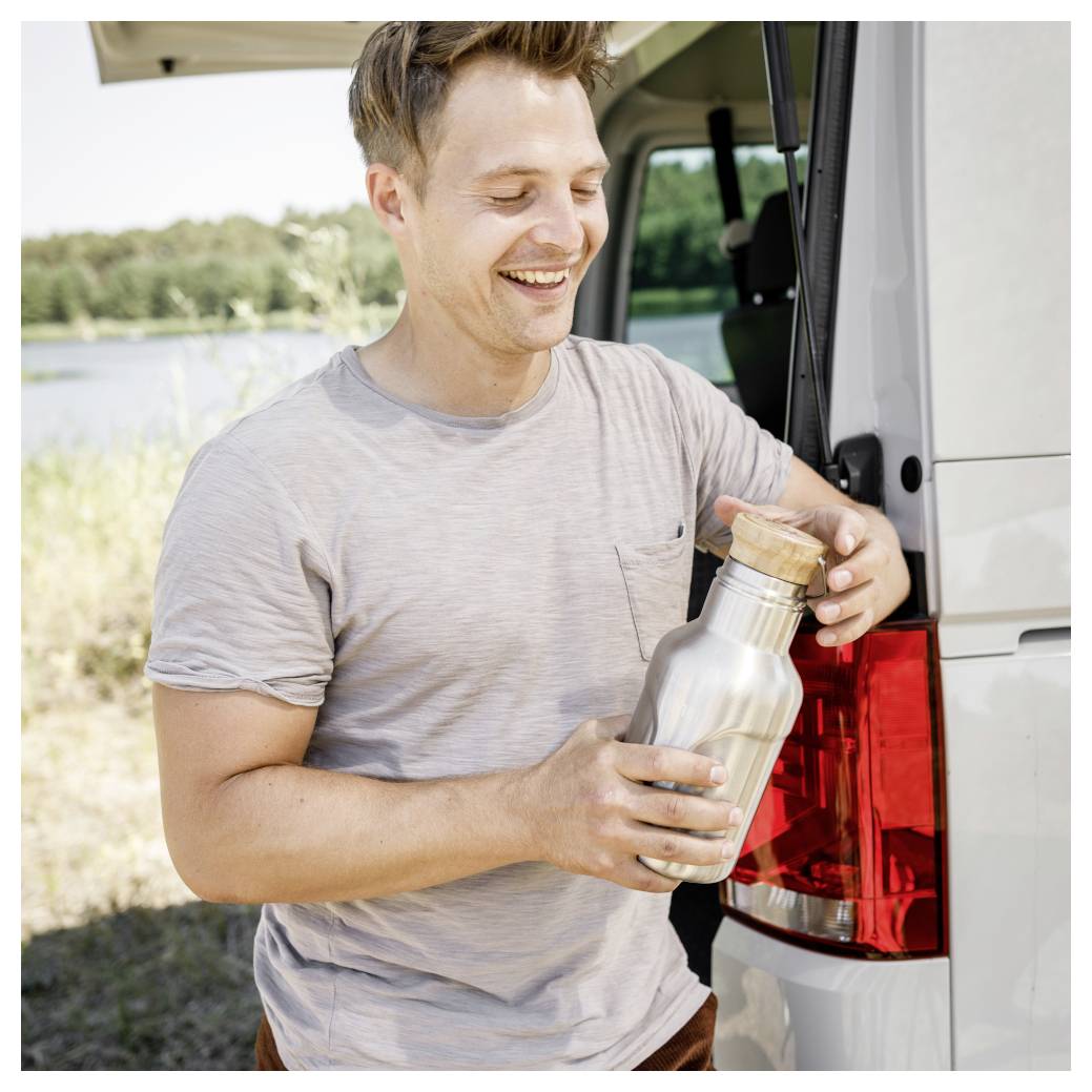 Ein lächelnder Mann neben einem Lieferwagen hält eine Edelstahl-Wasserflasche im Freien in der Nähe eines Sees, was auf eine Reise- oder Campingpause hindeutet.