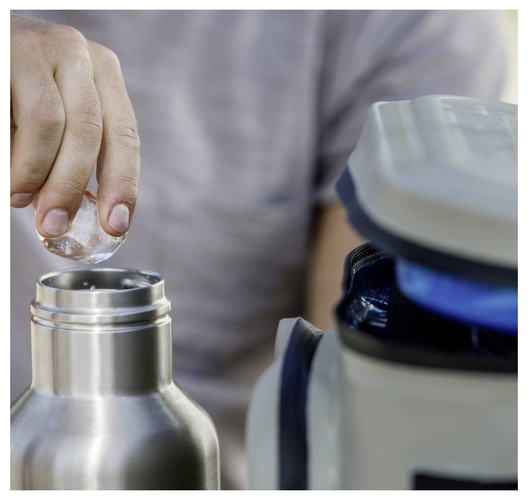 Eine Hand legt einen Eiswürfel in eine Edelstahlflasche neben einem Mixer auf einer Arbeitsplatte.