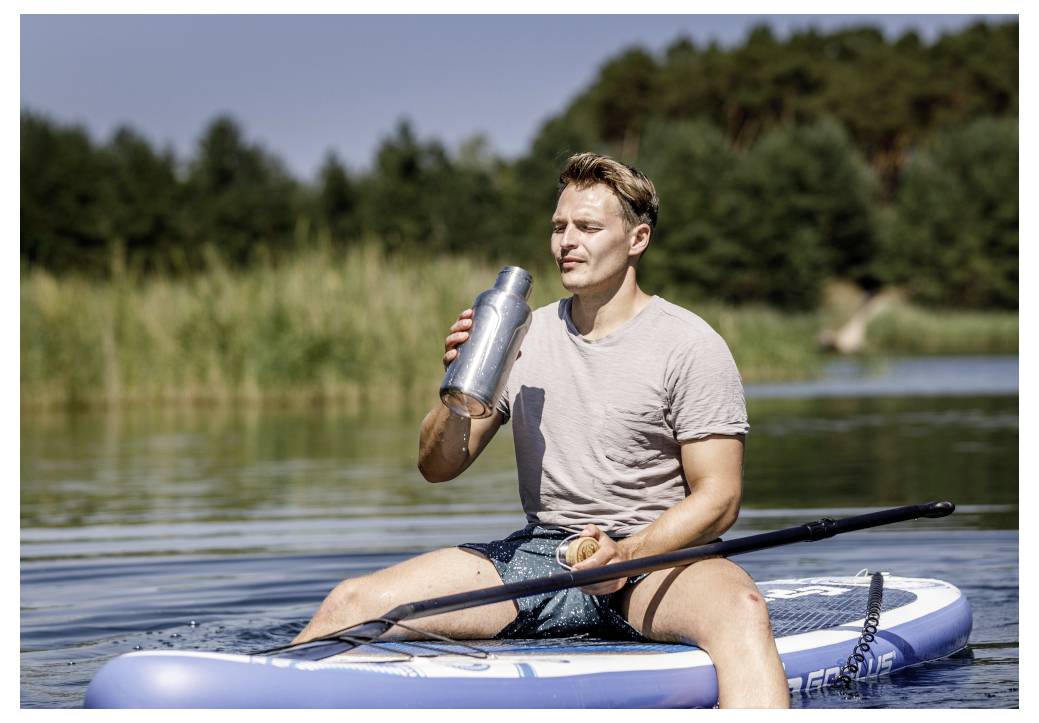 Ein Mann sitzt auf einem Paddleboard auf einem See, trinkt aus einer Metallwasserflasche und hält ein Paddel in der Hand.