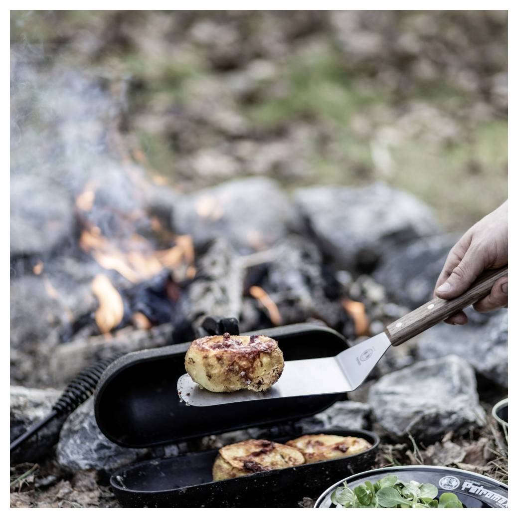 Eine Hand legt gegrillte Bratlinge in eine Pfanne über einem Lagerfeuer im Freien, mit Flammen und Steinen im Hintergrund.