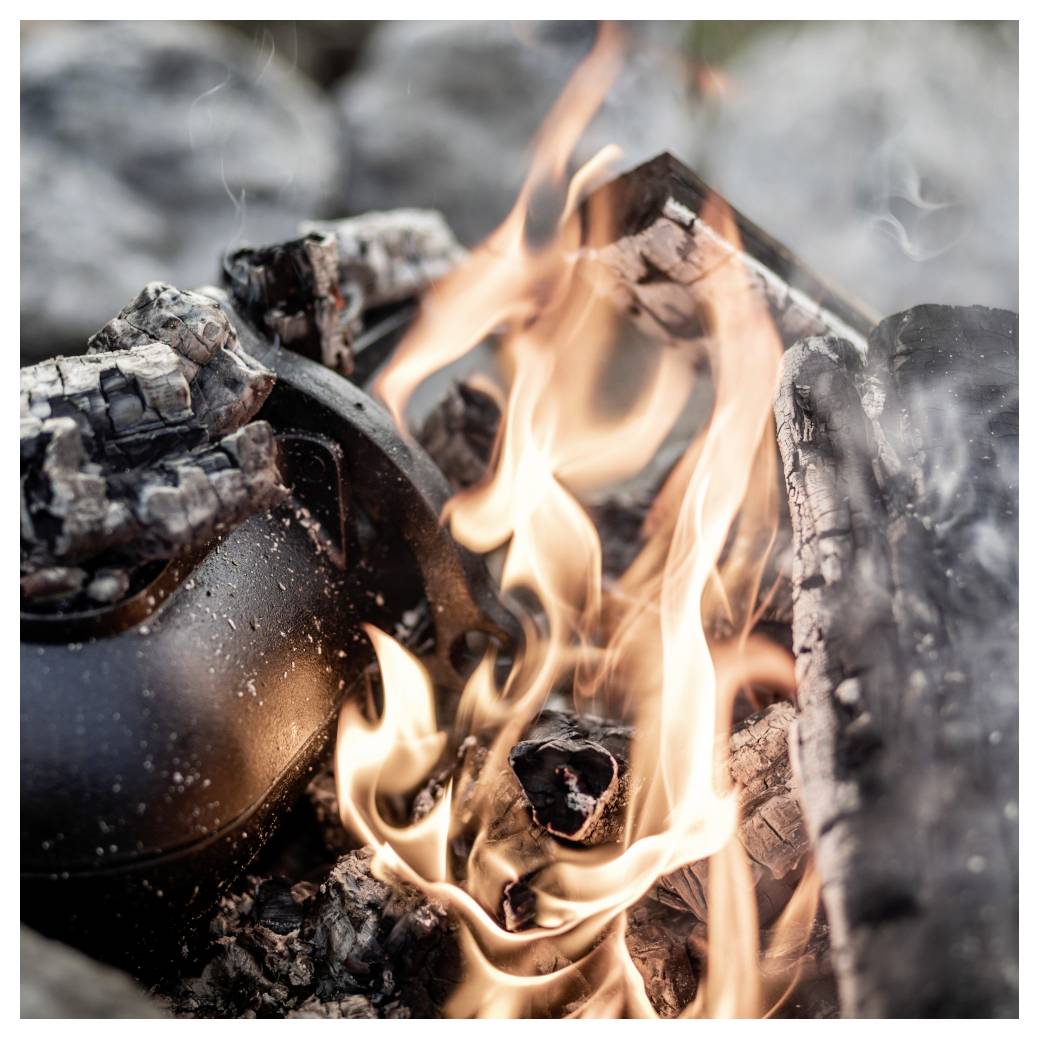 Schwarzer Kessel oder Topf, teilweise von brennenden Holzscheiten und Flammen in einem Lagerfeuer bedeckt, mit Rauch, der um ihn herum aufsteigt.