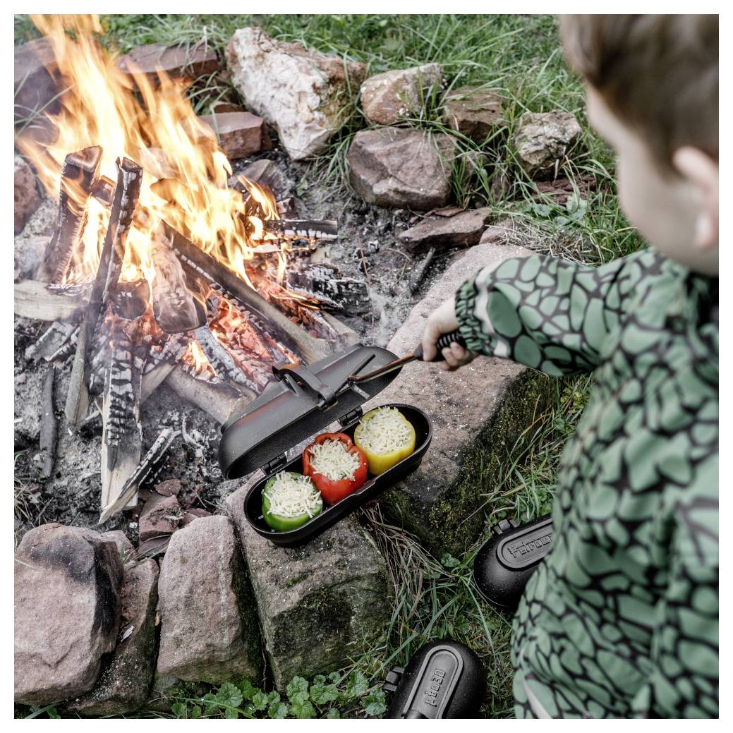 Kind am Lagerfeuer grillte gefüllte Paprika in einer tragbaren Pfanne neben Steinen und Gras, mit Kochutensilien in der Nähe.
