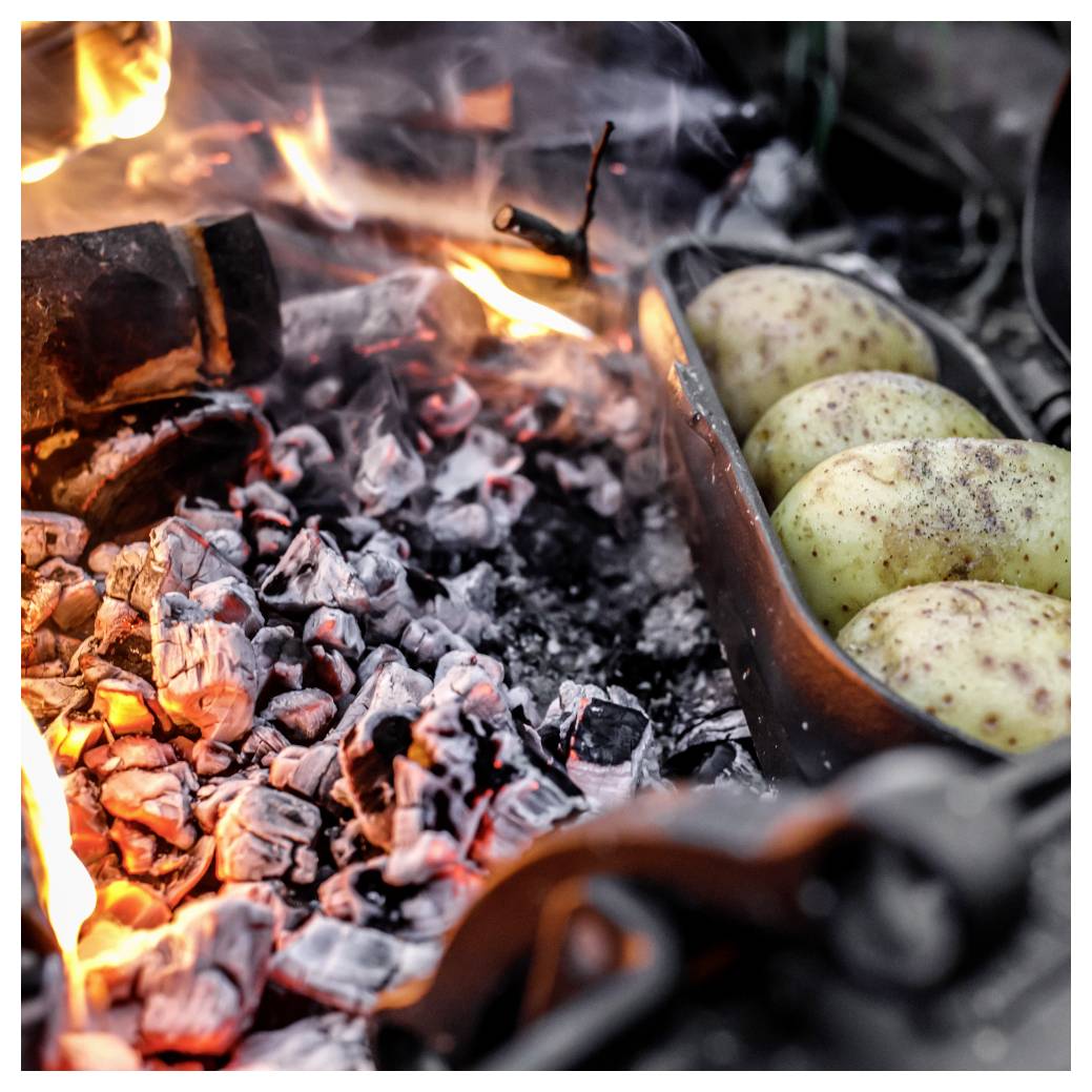 Kartoffeln rösten in einer Metallpfanne über heißen Lagerfeuerkohlen, mit Flammen und glühenden Gluten in der Nähe.