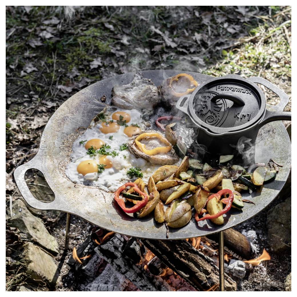 Eier, Kartoffeln, Gemüse und Fleisch, die auf einer gusseisernen Grillplatte über einem offenen Lagerfeuer im Freien garen.