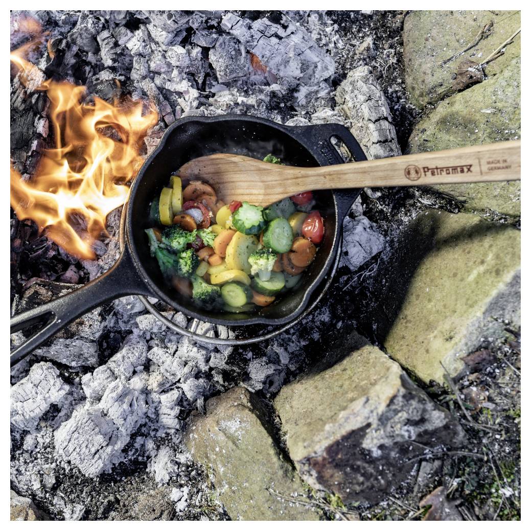 Gusseiserne Pfanne mit gemischtem Gemüse, die über einem offenen Lagerfeuer kocht, mit einem Holzspachtel, der über der Pfanne ruht.