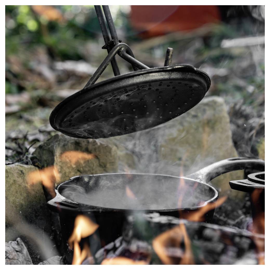 Gusseiserner Topf, der über einem offenen Lagerfeuer köchelt, mit einem darüber hängenden Deckel, Dampf und Flammen steigen draußen auf.