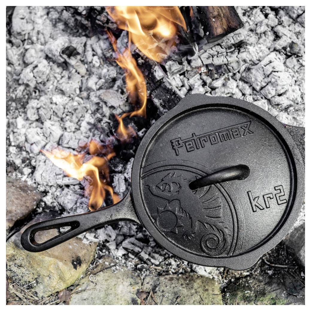 Gusseisen-Petromax-kr2-Pfanne mit Deckel, die in Lagerfeuer-Asche ruht, mit Flammen, die darum aufsteigen.