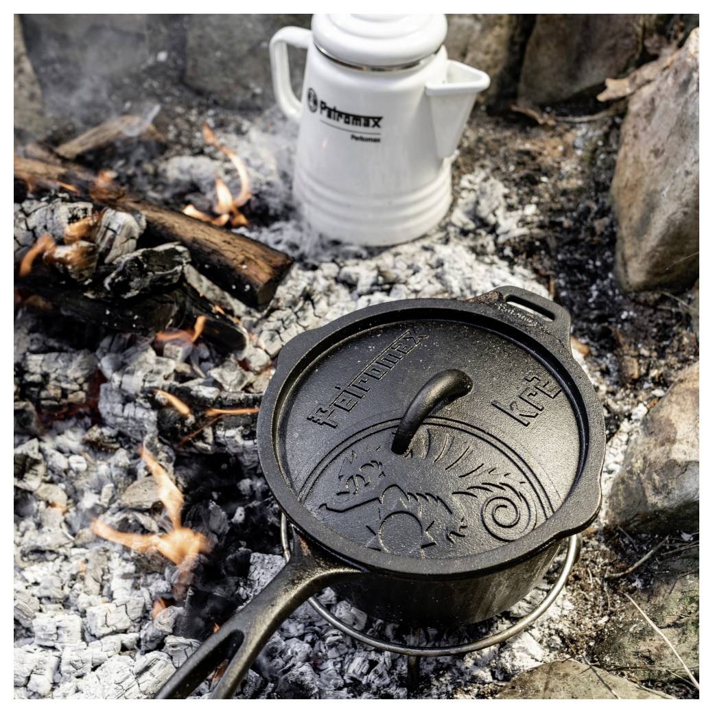 Gusseiserner Topf mit Deckel, der über einem Lagerfeuer auf glühenden Kohlen kocht, mit einem weißen Primus-Wasserkessel im Hintergrund.