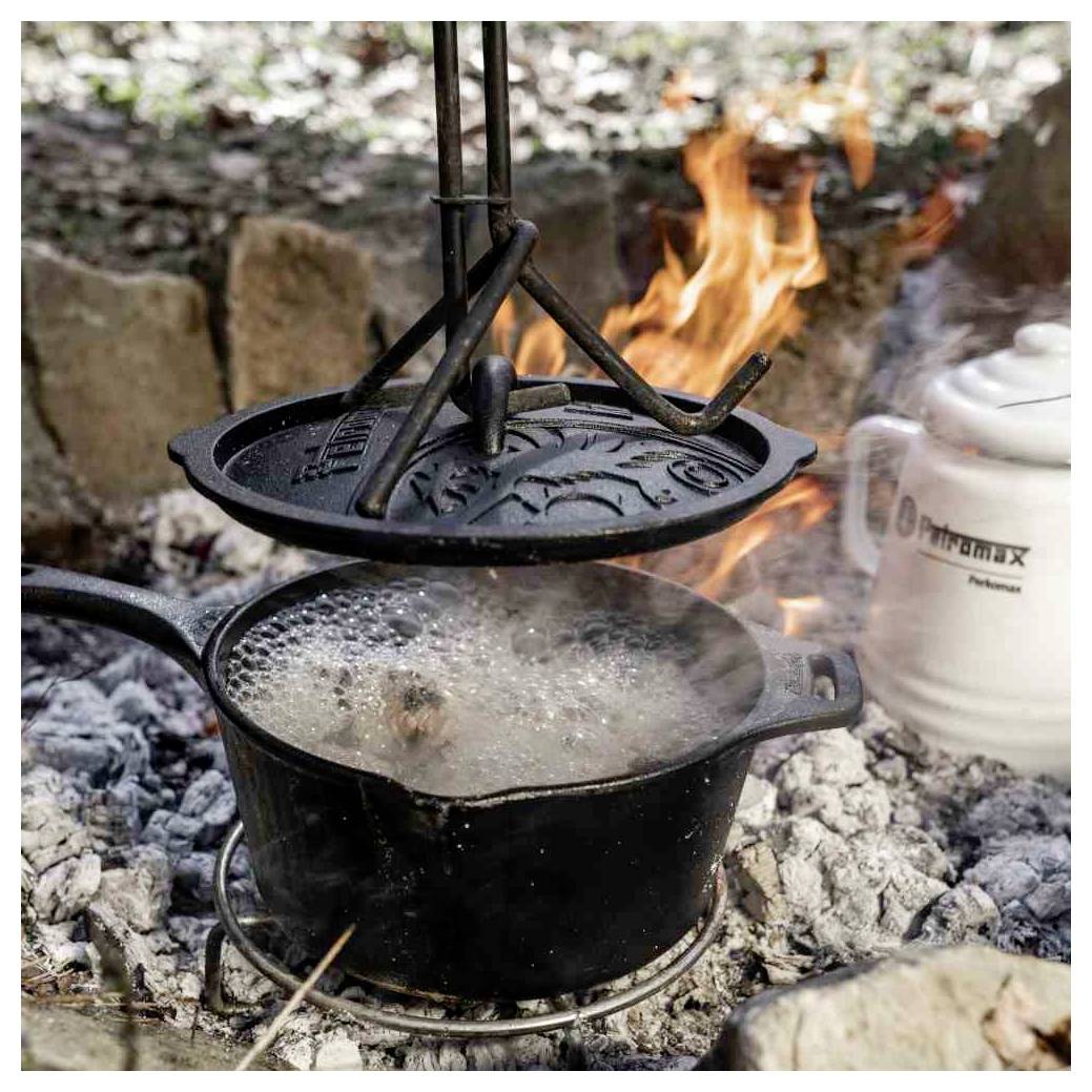 Ein Topf mit Essen kocht über einem offenen Lagerfeuer, mit einem Gusseisendeckel, der darüber hängt, und einem weißen Emailleekessel in der Nähe.