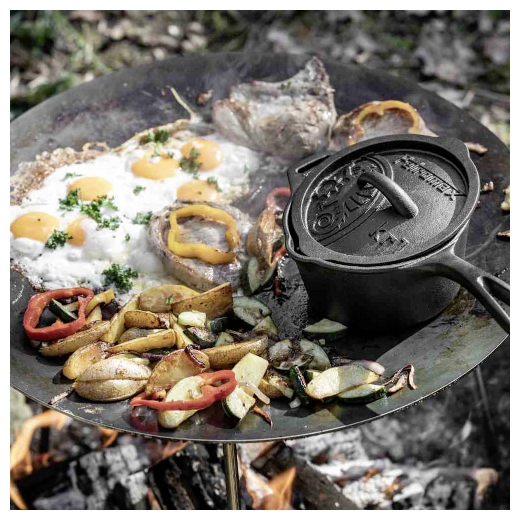 Gusseiserne Pfanne über einem Lagerfeuer mit Eiern, Kartoffeln, Gemüse, gegrilltem Fleisch und einem Deckel aus einem Dutch Oven, der im Freien kocht.