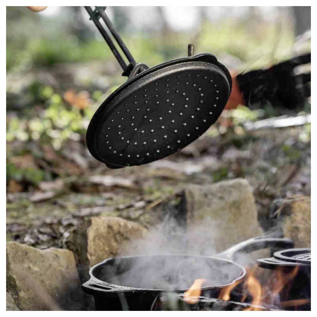 Ein gusseiserner Bräter, der über einem Lagerfeuer kocht, mit einem Deckel, der darüber hängt, Dampf steigt auf und Flammen sind zwischen den Steinen sichtbar.