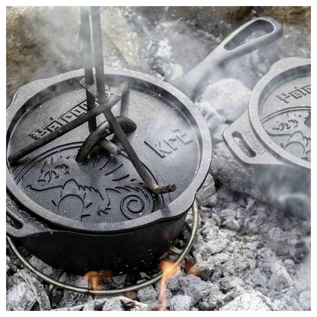 Gusseiserner Holländertopf beim Kochen über heißen Kohlen und Flammen, mit einer daneben ruhenden Deckelhebevorrichtung, im Freien