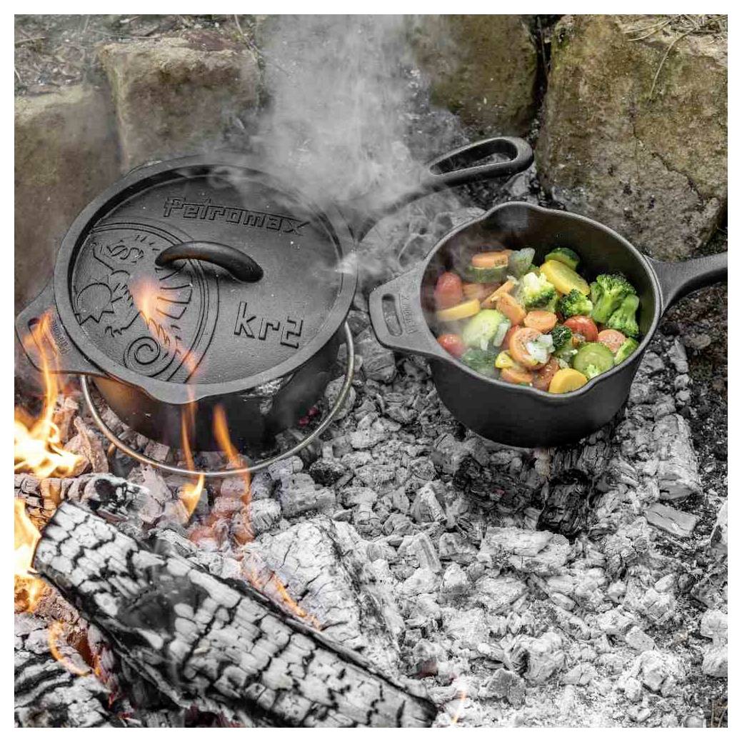 Gusseiserner Bräter und Bratpfanne, die über heißen Lagerfeuerkohlen kochen, mit Gemüse, das in der Pfanne schmort