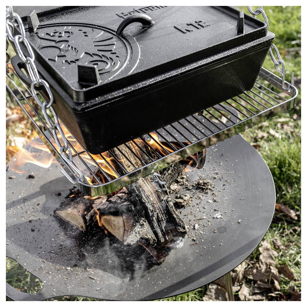 Gusseiserner Dutch Oven mit geprägtem Deckel hängt über einem Holzfeuer in einer runden Außenfeuerstelle und kocht über Flammen und Rauch.
