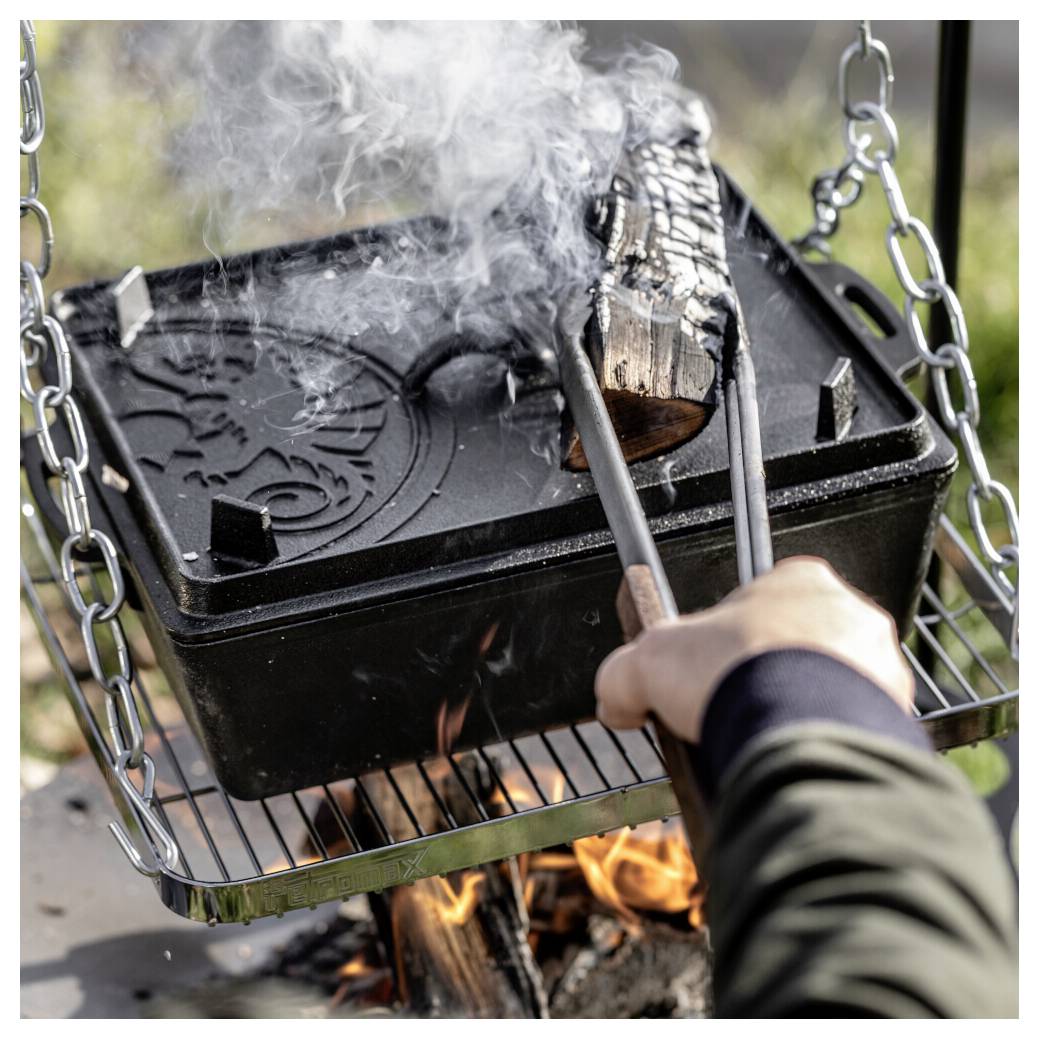 Person, die Essen in einem schwarzen gusseisernen Bräter über einem offenen Lagerfeuer grillt, Zangen verwendet, während Rauch aufsteigt.