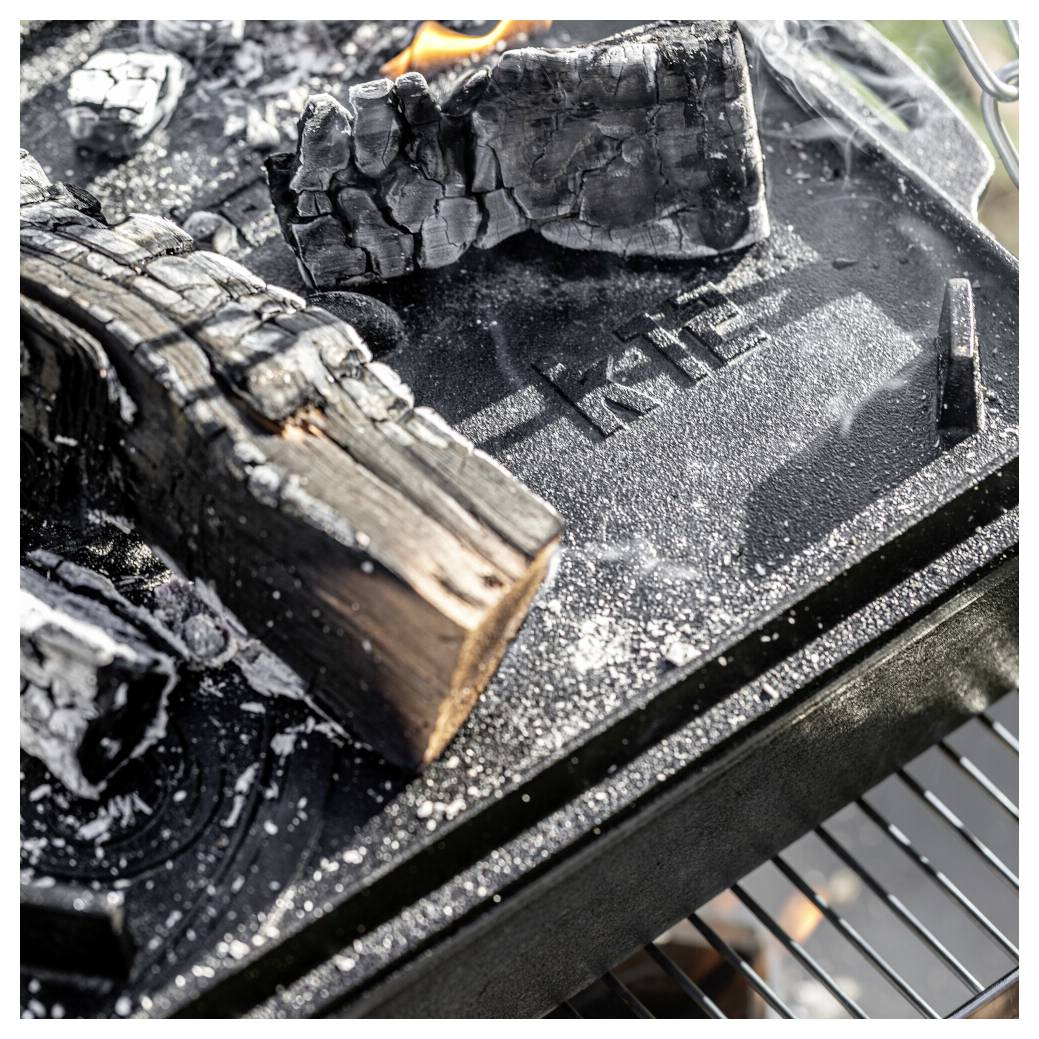 Verkohlte Holzscheite, die auf einem Grill über einer gusseisernen Platte oder einem Raucherblech brennen, mit sichtbaren Glut und Rauch.