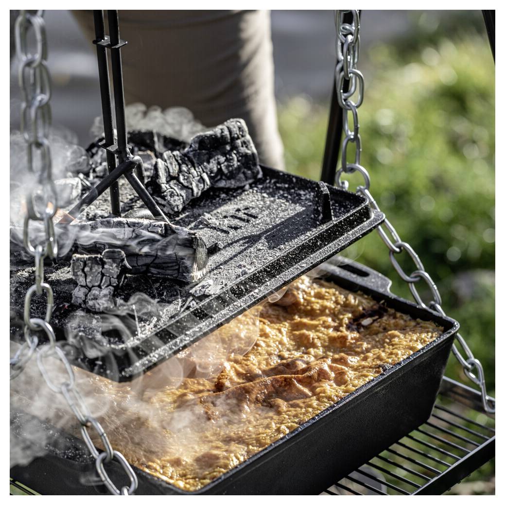 Ein gusseiserner Dutch Oven mit kochendem Eintopf, der über einem offenen Feuer kocht, mit heißen Kohlen auf dem Deckel und sichtbaren hängenden Kettenhalterungen.