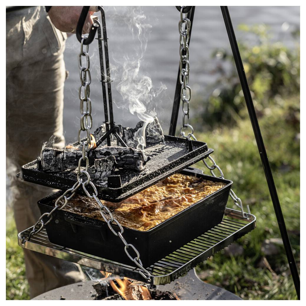 Eine Außen-Hängekocher-Einrichtung mit einer verkohlten oberen Pfanne, die über einem Feuer raucht, und einer unteren Pfanne, die darunter ein goldenes Auflaufformular backt.