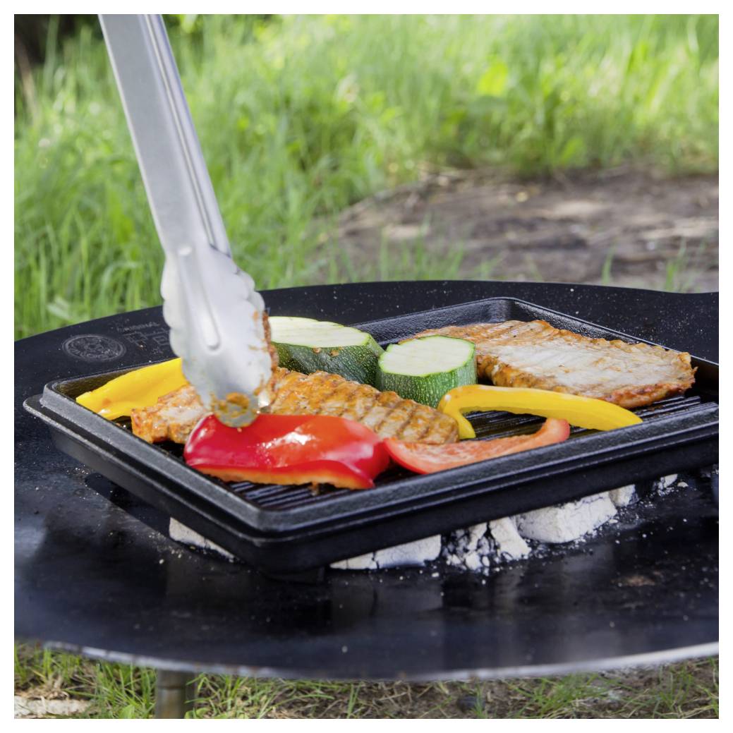 Außengrill mit einer Grillpfanne, die Gemüse und Fleisch zum Kochen hält, während Metalzangen ein Stück Essen über glühenden Kohlen umdrehen.