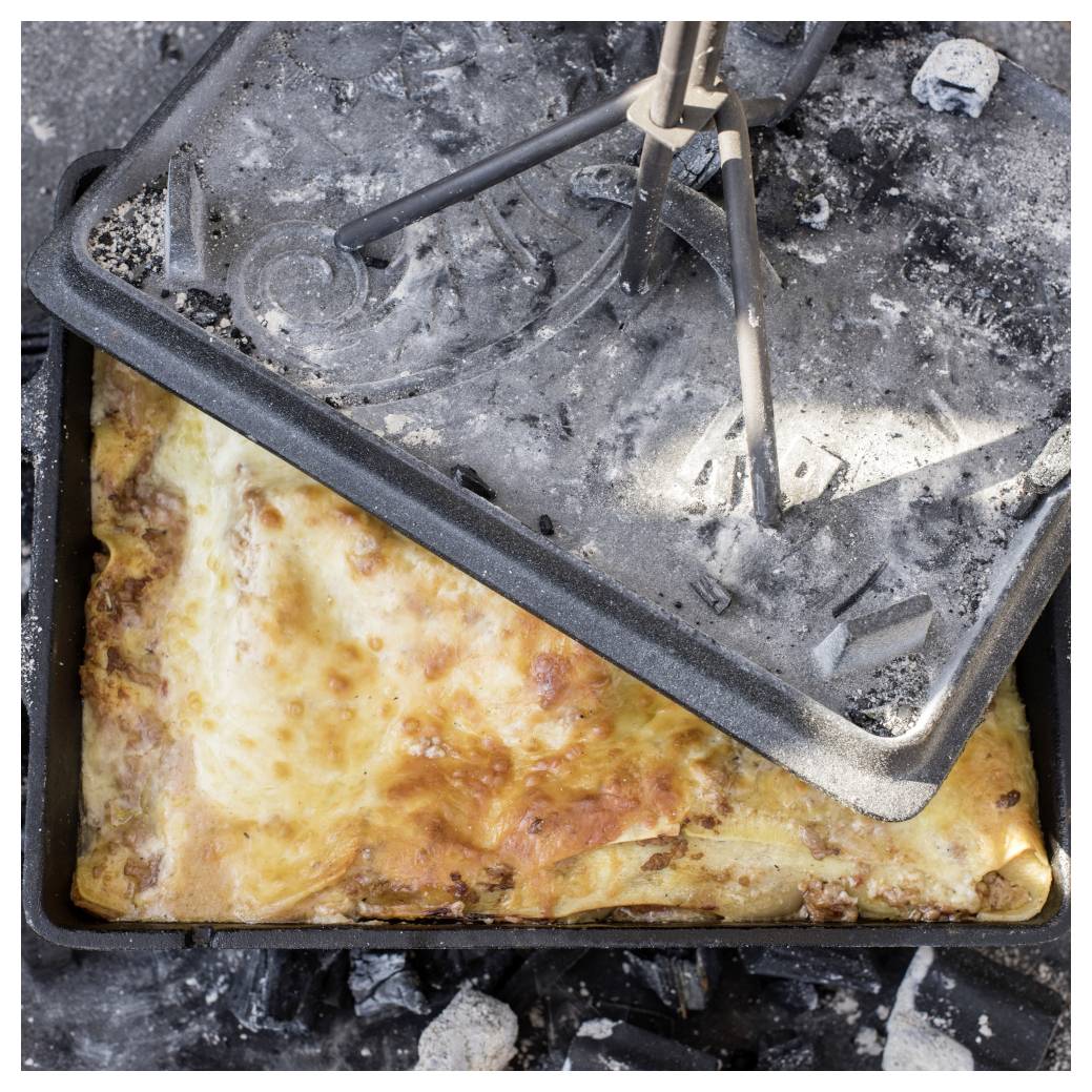 Offenes Lagerfeuer mit niederländischem Ofen mit goldbraun gebackenem Auflauf oder Brot innen, Deckel mit Asche bedeckt und mit einer Zange entfernt, Glut entfernt.