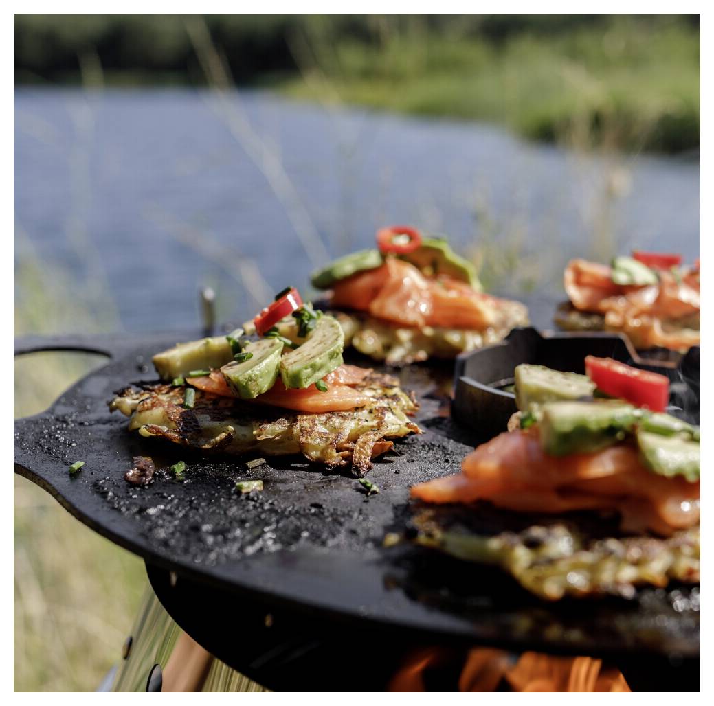 Nahaufnahme von gegrillten Gemüse- und Lachs-Patties, belegt mit Avocado und roten Chili-Pepperoni, die auf einem runden Outdoor-Grillrost am Wasser garen.