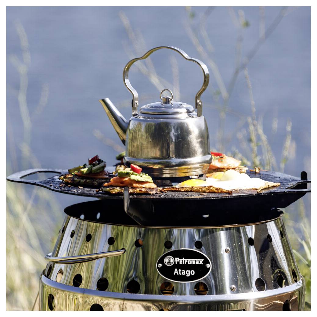 Edelstahlkessel auf einem runden Outdoor-Grill oder Ofen mit gekochtem Essen rundherum, vor einem unscharfen Wasserhintergrund.