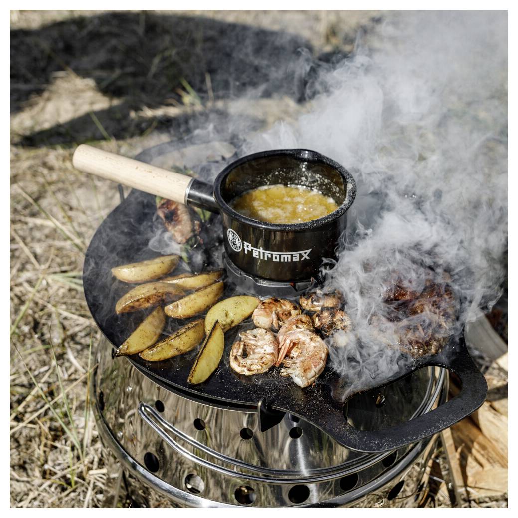 Outdoor-Lagerfeuer-Kochen auf einem tragbaren Kocher, mit einer Pfanne mit kochendem Essen und gegrillten Kartoffelecken und Fleisch, das inmitten von Rauch zischt.